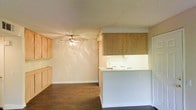 This single-floor space offers a practical and open layout that seamlessly connects the kitchen and living areas. The kitchen is equipped with light wood cabinetry that extends to the upper walls, providing ample storage space. A peninsula counter serves as a natural divider between the kitchen and the living area, creating a functional flow while maintaining an open feel. The countertops are finished in a light neutral tone that complements the warm wood tones of the cabinetry. A white range is visible within the kitchen, adding to the clean, understated aesthetic of the space. The living area is open and airy, featuring a ceiling fan with a light fixture that offers both ventilation and illumination. The flooring throughout is a dark hardwood or laminate, providing a grounding contrast to the light-colored walls and cabinetry. Neutral wall tones keep the space feeling bright and versatile, ready to accommodate a variety of interior styles. The overall ambiance is clean, functional, and welcoming, making it a practical space suited for everyday living.