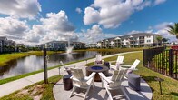 This single-story residence is set within a thoughtfully designed residential community that places a strong emphasis on outdoor living and scenic surroundings. The most notable feature of the property is its access to a beautifully landscaped outdoor common area positioned along a tranquil lake. A central fire pit, surrounded by white Adirondack chairs and cylindrical side tables, creates an inviting gathering space ideal for relaxation and socializing. The lake itself serves as a picturesque backdrop, highlighted by an elegant water fountain feature at its center. Manicured green lawns extend along the waterfront, and a paved pathway runs alongside the lake, offering a pleasant setting for walking. The multi-story residential buildings visible across the water feature classic architectural styling with neutral exteriors and private balconies. The overall ambiance of the outdoor environment is open and airy, benefiting from abundant natural light and expansive sky views. While interior imagery was not sufficiently captured to detail the indoor spaces, the community's outdoor amenities suggest a lifestyle-oriented environment designed for comfort and connection with nature.