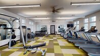 Located at 2785 Polo Club Blvd in Lexington, Kentucky, this single-floor fitness center is a comprehensively outfitted facility designed to support a wide range of workout routines. The space is divided into distinct functional zones, each tailored to a specific type of exercise.

The cardio area features a row of modern treadmills and elliptical machines positioned along windowed walls, allowing natural light to fill the space while providing an open, airy atmosphere. Overhead linear lighting and ceiling fans ensure consistent comfort throughout.

The strength training zone is equipped with a variety of cable and resistance machines, a dedicated free weights section with a full dumbbell rack, adjustable benches, and large wall-mounted mirrors that enhance the sense of space and allow users to monitor their form.

A separate functional training area features rubber mat flooring, spin bikes, medicine balls, stability balls, and a standing punching bag, offering a versatile space for high-intensity and group-style workouts. A wall-mounted TV is also present in this zone.

Throughout the facility, the flooring transitions between a distinctive green, tan, and brown patchwork tile in the main areas and dark rubber matting in the functional training zone. The white walls, abundant windows with shutters, and ceiling fans contribute to a clean, well-ventilated environment suited for focused, effective training sessions.