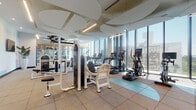 Located along Biscayne Boulevard in Miami's vibrant MiMo District, this contemporary fitness center is designed to deliver a premium workout experience. The space is defined by expansive floor-to-ceiling glass walls that flood the interior with abundant natural light and frame sweeping views of Miami's skyline and lush tree canopy, creating an energizing atmosphere throughout.

The main fitness floor features a comprehensive selection of equipment, including treadmills, elliptical trainers, stationary and Echelon bikes, and a rowing machine positioned along the window-facing wall to maximize the outdoor views during cardio sessions. A dedicated strength training area offers a full rack of dumbbells, kettlebells, weight benches, and a range of selectorized resistance machines. Full-length mirrors visually expand the space and support proper form during training.

A separate, glass-enclosed Training Zone provides a dedicated space for functional fitness, complete with hardwood flooring, wall-mounted storage racks, medicine balls, and resistance accessories — ideal for group or individual training sessions. The zone features a warm wood-slat ceiling that adds a distinct, boutique-gym feel.

The facility's design is anchored by sculptural, cloud-shaped ceiling panels throughout the main area, complemented by sleek linear lighting. Accent walls with geometric pastel murals add a modern aesthetic, while water fountains and mounted displays enhance convenience and comfort.