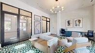 Located in Apopka, Florida, this thoughtfully designed amenity space combines functionality with contemporary style across a single floor. The main lounge area serves as the heart of the space, commanding attention with its striking green-and-white geometric patterned tile flooring, soaring white ceilings, and an elegant circular chandelier with globe bulbs that anchors the room. Large black-framed glass doors and transom windows flood the interior with natural light while offering views to the outdoors, creating an inviting, open atmosphere. Comfortable seating arrangements — including a striped sectional sofa, modern accent chairs, and a round coffee table with a potted plant — encourage collaboration and relaxation. Curated abstract and nature-inspired artwork adorns the white walls throughout, adding a polished, gallery-like quality to the space. A built-in teal credenza with task seating integrates a functional workspace seamlessly into the lounge. Two private office spaces provide quiet, focused work environments, each featuring a full-wall blue geometric wallcovering, a built-in floating desk, an ergonomic chair, plush carpet flooring, and framed bird-in-flight photography. Together, these offices offer a refined and productive retreat within an already well-appointed space.