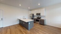 This single-floor unit at 3900 W Broad St in Richmond, Virginia offers a thoughtfully designed, contemporary living space with modern finishes throughout. The open-concept layout seamlessly connects the kitchen and living area, creating a spacious, cohesive environment ideal for everyday living and entertaining.

The kitchen is a standout feature, equipped with stainless steel appliances including a range, over-the-range microwave, and refrigerator. A large center island with a white quartz countertop and integrated sink provides ample workspace and additional seating. Two-tone cabinetry — white upper cabinets paired with dark lower cabinets — adds a modern aesthetic, while a herringbone tile backsplash introduces texture and visual interest. Warm wood-look flooring runs throughout the main living areas, contributing to a clean, contemporary feel.

The open living area is bright and generously proportioned, with recessed lighting and multiple windows that invite natural light and offer views of the surrounding neighborhood. The bedroom is a well-sized, quiet retreat featuring the same wood-look flooring and large windows with blinds. The bathroom includes a full tub-shower combination, tile flooring, and brushed gold hardware accents, lending a refined, modern touch to the space.