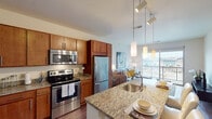 This thoughtfully designed single-floor residence offers a seamless blend of modern finishes and functional living spaces. The open-concept kitchen is a standout feature, equipped with stainless steel appliances including a range, over-the-range microwave, and refrigerator. Rich warm-toned wood cabinetry pairs beautifully with granite countertops, while a kitchen island with a built-in sink provides additional prep space and casual seating. Dark hardwood flooring runs throughout the kitchen and living areas, adding warmth and continuity. A built-in desk area within the kitchen offers a convenient workspace. The living room is bright and welcoming, anchored by large sliding glass doors that open onto a private balcony, inviting an abundance of natural light and offering sweeping views of the surrounding urban landscape. The bedroom is generously sized with plush carpeting, large windows framing city views, and ample storage. The bathroom features granite countertops, wood cabinetry, and a clean, neutral palette with tile flooring. Throughout the residence, the design emphasizes clean lines, quality materials, and a cohesive aesthetic that creates a comfortable and contemporary living environment.