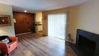 This single-floor residence offers a welcoming and functional layout designed for comfortable everyday living. The main living area is anchored by rich, wide-plank hardwood flooring that runs throughout, adding warmth and a sense of continuity to the space. A bold terracotta accent wall provides a striking focal point, lending character and depth to the room's aesthetic.

Natural light pours in through a large sliding glass door fitted with vertical blinds, creating a bright and airy atmosphere while offering direct access to the outdoors. The open layout allows the living area to flow seamlessly toward the kitchen, which is visible from the main room and features classic cream-colored cabinetry, dark countertops, and tile flooring that provides a clean contrast to the wood in the living space.

The overall ambiance is cozy yet open, with a neutral color palette accented by warm tones throughout. Decorative touches — including wall-mounted art and unique sculptural pieces — contribute to the space's lived-in, personalized character. The single-floor design ensures easy navigation and accessibility, making this a practical and inviting home.