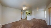 This single-floor residence offers a functional and straightforward layout connecting the main living spaces efficiently. The living room is a generously sized, open area covered in wall-to-wall carpet and illuminated by a ceiling fan with integrated lighting. It flows directly toward the kitchen, which is separated by a white peninsula counter — a practical divide that maintains an open feel between the two spaces. The kitchen is equipped with light wood cabinetry, white countertops, a coil electric range, an over-the-range microwave, and a full-size refrigerator. Hardwood-look flooring runs through the kitchen area, contrasting the carpeted living spaces.

The bedrooms continue the carpeted flooring and are each fitted with ceiling fans. One bedroom features a large sliding glass door with vertical blinds alongside a window overlooking a green outdoor area, allowing for abundant natural light. A second bedroom includes a walk-in closet and a side window. The bathroom is finished with wood-look plank flooring, a full tub/shower combination, a toilet, and a vanity with light wood cabinetry above. Overall, the unit presents a clean, practical interior with a neutral color palette throughout.