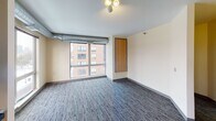 This single-floor unit offers a functional and thoughtfully designed layout ideal for comfortable urban living. The living area is finished with patterned blue-gray carpet and warm beige walls, creating a neutral, inviting atmosphere. Large windows allow generous natural light to fill the space while framing views of the surrounding cityscape and neighboring brick buildings. Exposed metallic ductwork runs along the ceilings, lending an industrial character throughout.

The kitchen is well-equipped with light maple cabinetry offering ample storage, a stainless steel double sink, and a full suite of black appliances including a refrigerator, electric range, microwave, and dishwasher. Light hardwood flooring in the kitchen provides a warm contrast to the carpeted areas of the unit.

The bedrooms are spacious and carpeted, each featuring large windows with city views and the same exposed ductwork detail overhead. Natural light flows freely into each room, making them feel open and airy.

The bathroom is designed with accessibility in mind, featuring white subway tile walls, a mosaic tile floor, an open roll-in shower with grab bars, a pedestal sink with mirror, and additional grab bar support throughout. The space is clean, bright, and practical, reflecting a commitment to inclusive design throughout the unit.
