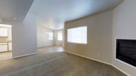 This single-story space presents an open-concept layout connecting a generously sized living area to an adjoining dining space. The living area is finished with wall-to-wall neutral-toned carpeting that extends throughout, complemented by smooth white ceilings and warm beige walls that create a cohesive, understated aesthetic. A large window fitted with horizontal blinds on the main wall allows natural light to filter into the living space, contributing to a bright and airy atmosphere. The transition into the dining area is seamless, where an elegant chandelier serves as a focal point overhead, adding a refined touch to the space. Additional windows in the dining area further enhance the natural light, creating a welcoming environment. Clean white baseboards frame the perimeter of both areas, providing a crisp contrast to the warm wall tones. The open flow between the two areas makes the overall layout feel expansive and functional, well-suited for both everyday living and entertaining. The neutral color palette throughout provides a versatile backdrop adaptable to a variety of interior design styles.