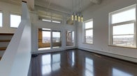 This single-floor space showcases an impressive open layout defined by its soaring double-height ceilings and crisp white walls, creating a bright and airy atmosphere. The main living area is anchored by rich, dark hardwood floors that reflect abundant natural light, adding warmth and depth to the space.

A prominent bank of large windows and glass sliding doors dominates one wall, framing sweeping views of the surrounding urban landscape, including exposed brick architecture typical of a historic city setting. Transom windows positioned above the main window line further enhance natural light penetration throughout the day. An additional window on the adjacent wall ensures the space remains well-lit from multiple angles.

A striking cluster of modern cylindrical pendant lights descends from the high ceiling, serving as both a functional and decorative centerpiece. Structural beams are visible overhead, adding an industrial character that complements the loft-like aesthetic of the space.

The sliding glass doors lead to an outdoor terrace area bordered by a low brick wall, extending the living space and offering direct access to outdoor views. Baseboard heating runs along the perimeter, ensuring comfort throughout the seasons. The overall ambiance is modern, open, and urban.