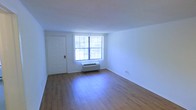 This single-floor space offers a clean, straightforward layout designed for comfortable everyday living. The main living area is finished with warm hardwood-style flooring that contrasts neatly against crisp white walls, creating a bright and welcoming atmosphere. A large window with vertical blinds serves as the focal point of the room, allowing generous amounts of natural light to fill the space throughout the day. A wall-unit HVAC system situated beneath the window provides both heating and cooling convenience directly within the living area. The entry door is positioned to one side, offering a practical and accessible point of entry. The textured white ceiling adds a subtle depth to the room's finish. The open nature of the main area provides flexibility for a variety of furniture arrangements and uses. Overall, the space presents a well-maintained, move-in ready environment with a neutral palette that makes it easy to personalize. Its simple yet functional design makes it well-suited for those seeking an efficient and comfortable living arrangement.