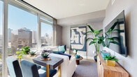 Located on the 15th floor at 110 Beech Street in downtown San Diego, this one-bedroom, one-bathroom residence offers an efficiently designed layout with modern finishes throughout. The living area is anchored by expansive floor-to-ceiling windows that flood the space with natural light and frame impressive panoramic views of the San Diego skyline. Warm hardwood-style flooring runs through the living and bedroom areas, complementing the neutral gray tones on the walls. The open-concept living and dining area flows seamlessly, making smart use of the available footprint. A built-in media console adds functional storage, while the kitchen features white cabinetry and stainless steel appliances. The bedroom is a well-appointed retreat, featuring a large floor-to-ceiling mirror that amplifies the sense of space, built-in shelving, warm bedside lighting, and continued hardwood-style flooring. The bathroom is finished with wood-tone flooring, a tiled tub-shower combination, and a bold teal accent wall that adds a distinctive design touch. Throughout the unit, a cohesive blue and teal color palette creates a calm, coastal ambiance that reflects its vibrant San Diego surroundings.