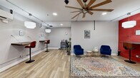 Located in the heart of Baltimore's Business Center (21201), this thoughtfully designed office space offers a versatile and welcoming environment ideal for professional use. The open single-room layout is finished with light hardwood flooring and neutral gray walls, creating a clean, modern aesthetic throughout.

A standout feature is the bold red exposed brick accent wall, which adds character and visual warmth to the space. A large, statement ceiling fan with wide wooden blades serves as a functional focal point overhead, complemented by recessed lighting and pendant fixtures that ensure the space is well-lit throughout the day.

The layout is divided into distinct functional zones. A comfortable lounge area is anchored by a vibrant patterned area rug and features two plush navy blue velvet armchairs with gold-accented legs, arranged around a stylish side table — ideal for client meetings or informal gatherings. Along the walls, wall-mounted bar-height desks with red tulip-style stools provide additional workspace options.

A black-framed glass-paned interior door adds an elegant, industrial touch and connects to an adjacent area. A beverage station and open shelving unit offer practical amenities for daily use. A wall-mounted split A/C unit ensures year-round comfort. The space strikes a confident balance between professional functionality and inviting design.