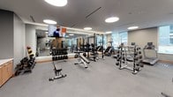 This single-level fitness center is thoughtfully designed to accommodate a full range of workout needs within a modern, upscale environment. The space is divided into distinct functional zones, each equipped with professional-grade machines and free weights. The strength training area features a comprehensive dumbbell rack, adjustable benches, a multi-station cable machine, and a power rack, all arranged on durable large-format gray tile flooring. Full-length mirrors line the walls, visually expanding the space and allowing users to monitor their form during exercises. A wall-mounted flat-screen television adds an entertainment element to the strength zone.

The cardio area is lined with a row of treadmills, elliptical trainers, and stair climbers, each equipped with individual digital screens. Floor-to-ceiling windows along one side of the cardio zone flood the space with natural light and offer views of the surrounding exterior. Warm wood-paneled accent columns provide an architectural contrast to the otherwise neutral palette of gray and white tones throughout.

Circular recessed lighting fixtures in the ceiling provide even, consistent illumination across the entire space. A glass-walled room visible from the main floor suggests an additional dedicated studio or stretching area, further enhancing the facility's versatility.