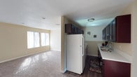 This single-floor space features a straightforward, functional layout that connects the living area and kitchen in an open, accessible arrangement. The living area is finished with wall-to-wall carpet and benefits from natural light streaming through a wide set of windows along one wall, creating a bright and comfortable atmosphere. Neutral-toned walls complement the light carpeting throughout this area. The kitchen is separated by a partial wall and transitions to dark hardwood-style flooring, offering a clear visual distinction between the two spaces. It is equipped with dark espresso-toned cabinetry, including upper cabinets that extend above the refrigerator for added storage. A full-size white refrigerator anchors the kitchen, accompanied by additional appliances and light-colored countertops that contrast with the darker cabinetry. A decorative area rug adds a touch of warmth to the kitchen floor. Both areas feature recessed and flush-mount ceiling lighting, and the textured ceiling is consistent throughout the space. Overall, the layout is practical and well-organized, with each area clearly defined while maintaining an open, connected feel.