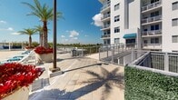 Located at 2500 SW 22nd Terrace in Fort Lauderdale, Florida, this outdoor amenity space presents a well-appointed resort-style pool deck designed for leisure and relaxation in a sunny South Florida setting. The centerpiece of the area is a generously sized swimming pool with clear, blue-tinted water, flanked on multiple sides by a broad, light-toned paver deck that provides ample room for movement and socializing. Rows of lounge chairs are arranged along the poolside, offering dedicated spaces for sunbathing and outdoor relaxation. Tall, mature palm trees are strategically positioned throughout the deck, lending a tropical character to the environment while providing natural shade. Vibrant red flowering plants in large planters add striking color accents against the crisp white façade of the multi-story residential building that frames the space. Shaded seating areas with umbrellas and cabana-style structures are available at the periphery, offering relief from the sun. Amenity structures including restroom facilities and utility access points are neatly integrated into the layout. A railed walkway connects the pool area to surrounding building access points, ensuring ease of circulation. Open views of the surrounding Fort Lauderdale skyline and lush greenery are visible beyond the perimeter, enhancing the overall outdoor experience. The deck is finished with large-format pavers that complement the clean, modern architectural aesthetic of the building.