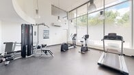 Located at 360 Torrance St in Burlington, Ontario, this fitness facility offers a compact yet well-appointed exercise room designed to support a well-rounded workout routine. The space is defined by its impressive floor-to-ceiling windows along one wall, which flood the interior with abundant natural light and provide open views of the surrounding greenery, creating an energizing atmosphere for exercise.

The room features durable rubber flooring in a speckled finish, ideal for high-traffic fitness use. Soaring ceilings with stylish pendant lighting add a sense of openness and volume to the space. The crisp white walls and clean, minimalist aesthetic contribute to a bright and welcoming environment.

Equipment includes a treadmill, elliptical trainer, stationary recumbent bike, rowing machine, a multi-function cable and weight machine, and a leg press/extension bench, along with free weights — offering a comprehensive range of cardio and strength training options. A wall-mounted split air conditioning unit ensures year-round climate comfort. Exercise reference charts are posted on the walls for user guidance. The space also includes a water cooler and recycling station near the exit, promoting convenience and sustainability. Overall, this fitness room delivers a functional and pleasant environment for residents or users seeking an accessible on-site workout facility.