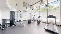 Located at 360 Torrance St in Burlington, Ontario, this fitness facility offers a compact yet well-appointed exercise room designed to support a well-rounded workout routine. The space is defined by its impressive floor-to-ceiling windows along one wall, which flood the interior with abundant natural light and provide open views of the surrounding greenery, creating an energizing atmosphere for exercise.

The room features durable rubber flooring in a speckled finish, ideal for high-traffic fitness use. Soaring ceilings with stylish pendant lighting add a sense of openness and volume to the space. The crisp white walls and clean, minimalist aesthetic contribute to a bright and welcoming environment.

Equipment includes a treadmill, elliptical trainer, stationary recumbent bike, rowing machine, a multi-function cable and weight machine, and a leg press/extension bench, along with free weights — offering a comprehensive range of cardio and strength training options. A wall-mounted split air conditioning unit ensures year-round climate comfort. Exercise reference charts are posted on the walls for user guidance. The space also includes a water cooler and recycling station near the exit, promoting convenience and sustainability. Overall, this fitness room delivers a functional and pleasant environment for residents or users seeking an accessible on-site workout facility.