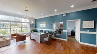 This single-floor leasing office is thoughtfully designed to create a professional yet inviting atmosphere for visitors. The space is defined by rich hardwood flooring that flows throughout, crisp white crown molding, and a striking teal accent wall that lends the interior a modern, coastal-inspired character.

Upon entry, guests are welcomed by a bright foyer featuring a cushioned bench, framed artwork, and glass-paneled double doors that invite natural light indoors. A pass-through window connects the entry to the main workspace, ensuring visibility and easy communication across the office.

The central work area is anchored by a contemporary light wood reception desk, accompanied by guest seating and additional workstations. Recessed ceiling lights and a stylish geometric chandelier provide warm, layered illumination. A large mirror with a gold frame adds depth and elegance to the teal feature wall.

A dedicated lounge area offers a comfortable retreat with plush cognac leather barrel chairs, a stone-topped coffee table, and an expansive picture window that frames lush outdoor greenery and community views, flooding the space with natural light.

An adjacent private office provides a quieter setting for consultations, while a utility corridor with louvered doors adds functional storage. Throughout, the cohesive color palette of soft grey, white, and teal creates a calm, professional environment well-suited for a leasing or administrative office.