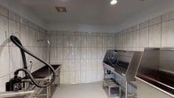 This single-floor professional pet grooming facility is purpose-built for efficiency and hygiene. The main grooming room features floor-to-ceiling large-format ceramic wall tiles in a neutral gray tone, creating a clean, clinical aesthetic that is easy to maintain. The smooth, light-colored flooring is designed for practicality, with a central floor drain ensuring efficient water management during bathing sessions.

The room is outfitted with multiple stainless steel dog bathing tubs, each elevated on step stools to allow easy access for pets and groomers alike. The tubs are equipped with integrated spray hose systems for thorough rinsing. Alongside the tubs, stainless steel grooming tables provide dedicated workstations, and wall-mounted professional dryer systems with flexible hoses are positioned for convenient use. Recessed ceiling lighting illuminates the workspace evenly, complemented by a ceiling-mounted HVAC vent for climate and air quality control.

The entry area transitions from the exterior through a full-height glass door with sidelights, allowing natural light to filter into the space and providing visibility to the parking area outside. The overall design of the facility prioritizes cleanliness, functionality, and a professional workflow suitable for high-volume pet grooming operations.