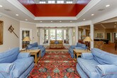 This single-floor space is designed for both relaxation and entertainment, offering a thoughtfully arranged layout with distinct areas that flow naturally into one another. The centerpiece of the space is a striking lounge area featuring soaring tray ceilings accented with bold red paneling, recessed lighting, and elegant crown molding. Comfortable blue slipcovered sofas are arranged around a vibrant red patterned area rug, while French doors and large windows invite abundant natural light and outdoor views. A wall-mounted television, a gold-framed mirror, and decorative wall accents add character to the room.

The expansive game room is a highlight of the space, showcasing warm terracotta brick flooring that extends throughout much of the floor plan. Exposed brick columns frame the room, lending a rustic yet refined aesthetic. A full-size pool table and a foosball table anchor the space, complemented by a well-appointed bar area with mirrored backing and seating along the counter. Adjacent to the game room, a dedicated dining area features sage green walls, recessed lighting, a large farmhouse-style wooden table with seating for eight, and a classic dark-wood display cabinet. The cohesive brick flooring and rich area rugs throughout tie the entire space together with warmth and visual continuity.