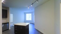 This single-floor space showcases a contemporary open-plan design that seamlessly integrates the kitchen and living areas. The kitchen features a dark-cabineted island with a light-toned countertop, providing both prep space and storage through multiple drawers and cabinet doors with sleek bar-style hardware. Overhead, a track lighting system runs along the ceiling, offering adjustable, directed illumination throughout the space. The flooring is a rich, dark hardwood that runs continuously through the open area, grounding the overall aesthetic and contrasting nicely with the crisp white walls and bright white baseboards. Natural light enters generously through a large window fitted with horizontal blinds, creating a bright and airy atmosphere in the main living area. A secondary window or door opening is visible nearby, further enhancing the sense of openness and ventilation. The walls are finished in a clean, neutral white, providing a versatile backdrop suitable for a variety of interior styles. The overall ambiance is modern, minimalist, and well-lit, making efficient use of the available floor space in a functional and visually cohesive layout.