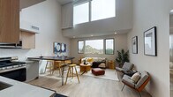 This two-story residence is defined by its striking architectural design, most notably the dramatic double-height ceilings in the main living area that create an expansive, airy atmosphere. Large black-framed windows on the main floor frame treetop views and draw in generous amounts of natural light, further enhancing the sense of openness.

The main floor integrates the living, dining, and kitchen areas in a cohesive open-concept layout. Light hardwood flooring flows throughout, complementing the neutral palette. The kitchen is well-appointed with light wood upper cabinetry, gray lower cabinets, stainless steel appliances including a gas range and dishwasher, and a white subway tile backsplash. A dark wood high-top dining table with hairpin legs and yellow bar stools provides a functional and stylish dining area adjacent to the kitchen.

The living area is generously proportioned, offering distinct seating zones beneath the soaring ceiling. A full bathroom on this level features a glass-enclosed walk-in shower with white tile surround, a wood-fronted vanity, and stone-look flooring.

The upper level hosts the bedroom, a calm and well-lit retreat with recessed lighting, plush carpeting, and a window overlooking the surrounding neighborhood. The overall ambiance is clean, contemporary, and inviting throughout.
