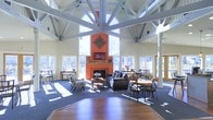 This impressive single-floor space is defined by its dramatic architectural character and open, airy atmosphere. The centerpiece of the room is a bold orange accent wall housing a classic fireplace, adorned with decorative elements and topped with a mounted television, creating a warm and inviting focal point. Above, an intricate radial white timber truss system fans out from a central hub beneath a soaring vaulted ceiling, lending the space a distinctive architectural identity. Expansive windows wrap around the perimeter, flooding the interior with generous natural light and offering views of the surrounding outdoor landscape. The flooring transitions between rich hardwood near the entry and plush dark carpeting in the seating areas, subtly delineating different zones within the open layout. The lounge area is furnished with leather sofas and patterned accent chairs arranged around the fireplace, encouraging relaxed conversation. Toward the perimeter, dining tables and cross-back chairs provide additional seating for gatherings. Sturdy structural columns frame the space without interrupting its openness. Overall, the space exudes a welcoming, communal ambiance that balances rustic warmth with refined architectural detail.