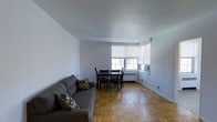 This single-floor space offers a clean and functional layout designed for comfortable everyday living. The combined living and dining area serves as the heart of the unit, characterized by warm parquet hardwood flooring arranged in a classic herringbone-style pattern that adds a timeless charm to the interior. Smooth, neutral-toned walls and a white ceiling create a bright and airy atmosphere throughout the space. Multiple windows along one wall allow a generous amount of natural light to filter in, enhancing the open feel of the room. A window-mounted air conditioning unit provides climate control. The living area comfortably accommodates a cushioned sofa, while the dining space features a modest table with seating for four, making the layout well-suited for both relaxing and entertaining. White roller shades on the windows offer adjustable privacy and light control. The space is finished with clean white baseboards that frame the flooring neatly. Overall, the unit presents a straightforward, well-lit environment with functional amenities and classic design elements that provide a solid foundation for personalization.