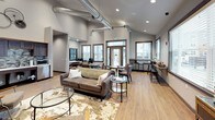 Located at 1608 East South Riverton Avenue in Spokane, WA, this single-floor community amenity space combines modern industrial design with warm, inviting finishes. The open-concept main lounge is anchored by soaring vaulted ceilings with exposed ductwork, recessed lighting, and large windows that fill the space with abundant natural light. A dramatic floor-to-ceiling natural stone fireplace serves as a focal point, complemented by a wall-mounted television and comfortable seating arranged for both relaxation and socializing. Light hardwood flooring runs throughout, enhancing the bright, airy atmosphere. The fully equipped kitchen features dark cabinetry, a mosaic tile backsplash, stainless steel appliances including a gas range, refrigerator, microwave, and dishwasher, along with ample counter space. A glass door leads directly to an outdoor patio area, offering convenient access to exterior amenities. Café-style counter seating along the windows and multiple dining tables provide a variety of gathering options. A connecting hallway, accented with curated artwork and a water fountain, leads to a dedicated fitness center. The overall design balances contemporary and industrial elements, creating a functional and welcoming shared space.