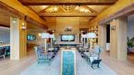 Located at 2080 FM 1102 in New Braunfels, Texas, this thoughtfully designed single-story clubhouse for the Landhaus community offers a generous and inviting gathering space defined by its distinctive Hill Country-inspired architecture. The interior is characterized by an abundance of natural wood — from wide-plank light hardwood floors to tongue-and-groove wood-clad walls and ceilings — creating a warm, cohesive aesthetic throughout.

At the heart of the space is a prominent reception area anchored by a stylish front desk, exposed reclaimed timber beams soaring overhead, and built-in shelving with curated displays. The grand living area features vaulted ceilings with dramatic wood beam trusses, a linear fireplace, and generous seating arrangements designed for both relaxation and conversation. A well-appointed mailroom with individual mailboxes is integrated into the lounge area.

Additional private rooms, including a cozy meeting or office space, branch off the main corridor. Floor-to-ceiling glass entry doors and large windows at multiple points flood the interior with natural light while offering views of the surrounding outdoor areas. Lush indoor greenery, layered lighting, and carefully selected furnishings complete this welcoming, resort-inspired community space.