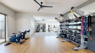 This fitness center in the 95356 area of Modesto, California, is a thoughtfully designed, single-story facility offering a comprehensive workout environment across multiple connected zones. The space features a clean, modern aesthetic defined by crisp white walls, recessed lighting, and ceiling fans that maintain comfortable airflow throughout.

The main cardio area is generously proportioned, with light-toned flooring underfoot and an array of equipment including treadmills, elliptical machines, stair climbers, and rowing machines. Full-length mirrors line the walls, visually expanding the space and supporting proper form during workouts. A dedicated cycling area features Matrix spin bikes positioned on plush carpeted flooring and is lined with floor-to-ceiling mirrors on multiple walls.

A separate strength training zone houses a multi-functional cable machine, a comprehensive dumbbell rack with a full weight range, kettlebells, and a Matrix adjustable bench, providing versatile options for resistance training. Large sliding glass doors in one section open to an outdoor terrace, bringing in abundant natural light and views of the surrounding community. Windows throughout the facility further brighten the interior, contributing to an open, airy atmosphere well-suited for focused exercise.