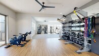 This fitness center in the 95356 area of Modesto, California, is a thoughtfully designed, single-story facility offering a comprehensive workout environment across multiple connected zones. The space features a clean, modern aesthetic defined by crisp white walls, recessed lighting, and ceiling fans that maintain comfortable airflow throughout.

The main cardio area is generously proportioned, with light-toned flooring underfoot and an array of equipment including treadmills, elliptical machines, stair climbers, and rowing machines. Full-length mirrors line the walls, visually expanding the space and supporting proper form during workouts. A dedicated cycling area features Matrix spin bikes positioned on plush carpeted flooring and is lined with floor-to-ceiling mirrors on multiple walls.

A separate strength training zone houses a multi-functional cable machine, a comprehensive dumbbell rack with a full weight range, kettlebells, and a Matrix adjustable bench, providing versatile options for resistance training. Large sliding glass doors in one section open to an outdoor terrace, bringing in abundant natural light and views of the surrounding community. Windows throughout the facility further brighten the interior, contributing to an open, airy atmosphere well-suited for focused exercise.