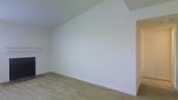 This single-story space presents a straightforward, functional interior characterized by its clean and understated aesthetic. The room showcased features smooth, white-painted walls that create a bright and neutral backdrop, complemented by white baseboards that provide a crisp finishing detail along the perimeter. The flooring consists of wall-to-wall carpeting in a muted beige tone, offering a soft and practical surface underfoot. The ceiling follows a slightly angled or stepped architectural line, adding a subtle structural dimension to the otherwise simple space. Electrical outlets are conveniently positioned along the walls, and a light switch is also visible, indicating the room is fully wired and functional. The overall palette is minimal and neutral, making the space highly versatile and move-in ready for a variety of uses. The absence of furnishings allows the full scale and layout of the room to be appreciated, revealing a generous open area suitable for flexible arrangement. The single-floor layout contributes to ease of accessibility throughout.