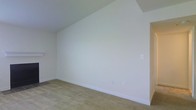 This single-story space presents a straightforward, functional interior characterized by its clean and understated aesthetic. The room showcased features smooth, white-painted walls that create a bright and neutral backdrop, complemented by white baseboards that provide a crisp finishing detail along the perimeter. The flooring consists of wall-to-wall carpeting in a muted beige tone, offering a soft and practical surface underfoot. The ceiling follows a slightly angled or stepped architectural line, adding a subtle structural dimension to the otherwise simple space. Electrical outlets are conveniently positioned along the walls, and a light switch is also visible, indicating the room is fully wired and functional. The overall palette is minimal and neutral, making the space highly versatile and move-in ready for a variety of uses. The absence of furnishings allows the full scale and layout of the room to be appreciated, revealing a generous open area suitable for flexible arrangement. The single-floor layout contributes to ease of accessibility throughout.