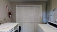 This dedicated laundry space is designed with functionality and organization in mind. The room's most prominent feature is a set of wide, white louvered bifold doors that span nearly the full width of the wall, offering ventilated access to a generous storage or utility closet behind them. The slatted design of the doors promotes airflow while maintaining a clean, uniform appearance.

Flanking the bifold doors on either side are white countertops that provide practical workspace for folding, sorting, or storing laundry essentials. A front-loading washer is positioned to the left beneath the counter, while a matching dryer unit occupies the right side, creating a symmetrical and efficient layout. The countertops extend over both appliances, maximizing usable surface area.

The walls are finished in a neutral warm gray tone, lending the room a calm, understated ambiance that complements the crisp white cabinetry and appliances. The overall design prioritizes utility without sacrificing a tidy aesthetic. This single-floor space is thoughtfully laid out to streamline laundry tasks while offering ample storage solutions, making it a highly functional area within the home.