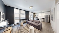 This compact, well-designed one-bedroom, one-bathroom unit offers a thoughtfully organized layout across a single floor. The entrance hallway, finished with wood-look flooring and clean white walls, leads directly into the open-plan living and kitchen area, establishing a modern, cohesive aesthetic from the moment you step inside.

The living area features a bold dark accent wall contrasted by large windows fitted with roller shades, allowing ample natural light to fill the space. Wood-look flooring continues throughout, complementing the contemporary furnishings. A sliding barn-style door separates the living area from the bedroom, adding a functional and stylish design element.

The kitchen is well-appointed with full-height white cabinetry, a white subway tile backsplash, white quartz countertops, and a complete suite of stainless steel appliances including a range, microwave, dishwasher, and refrigerator. Track lighting overhead enhances both functionality and ambiance. A compact dining table with seating sits adjacent to the kitchen.

The bedroom is an intimate, carpeted retreat featuring a ceiling fan, textured wallpaper accent, and warm bedside lighting. The bathroom maintains the unit's clean aesthetic with white cabinetry, a vanity with mirror, and wood-look tile flooring.