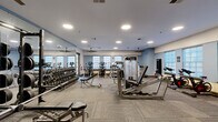 This fitness center offers a thoughtfully designed, single-floor exercise space that provides ample room for a wide variety of workouts. The interior features neutral-toned carpet tiles, white and light blue walls, and a bright, clean aesthetic reinforced by recessed circular ceiling lights and multiple ceiling fans that keep the environment comfortable during intense sessions.

Large windows line the perimeter walls, allowing natural light to fill the space and creating an open, energizing atmosphere. Full-wall mirrors along one side visually expand the room while allowing users to monitor their form during strength training.

The equipment selection is extensive and well-organized. A dedicated free weights area features a full dumbbell rack paired with flat benches, while a variety of selectorized strength machines — including cable systems, a leg press, and upper-body stations — are distributed throughout the floor. Cardio equipment is equally well-represented, with multiple treadmills equipped with individual display screens, stationary cycling bikes, elliptical trainers, and a stair climber. A freestanding punching bag rounds out the offerings for those interested in combat fitness training.

The layout promotes smooth circulation between zones, and glass-paned doors provide easy access to and from the space. Overall, this fitness center is a comprehensive, professional-grade facility suited for a full range of training goals.