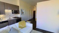 This single-floor space showcases a thoughtfully designed open-concept layout that seamlessly connects the living and kitchen areas. The kitchen is outfitted with dark, flat-panel cabinetry that provides a striking contrast against light-toned walls, complemented by stainless steel appliances including a gas range and built-in microwave. A classic subway-style mosaic backsplash adds texture and visual interest to the cooking space.

The living area flows naturally from the kitchen, featuring rich, dark hardwood flooring that runs consistently throughout the unit, contributing to a cohesive and polished aesthetic. A white sofa anchors the living space, accented by a plush area rug and a sleek mirrored coffee table, creating a clean and contemporary feel.

A hallway extending from the main living area leads to additional rooms, flanked by white paneled doors and overhead ventilation. Neutral gray walls maintain a calm, modern ambiance throughout, while recessed lighting ensures consistent illumination. The overall design balances functionality with a refined, urban aesthetic.