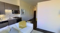 This single-floor space showcases a thoughtfully designed open-concept layout that seamlessly connects the living and kitchen areas. The kitchen is outfitted with dark, flat-panel cabinetry that provides a striking contrast against light-toned walls, complemented by stainless steel appliances including a gas range and built-in microwave. A classic subway-style mosaic backsplash adds texture and visual interest to the cooking space.

The living area flows naturally from the kitchen, featuring rich, dark hardwood flooring that runs consistently throughout the unit, contributing to a cohesive and polished aesthetic. A white sofa anchors the living space, accented by a plush area rug and a sleek mirrored coffee table, creating a clean and contemporary feel.

A hallway extending from the main living area leads to additional rooms, flanked by white paneled doors and overhead ventilation. Neutral gray walls maintain a calm, modern ambiance throughout, while recessed lighting ensures consistent illumination. The overall design balances functionality with a refined, urban aesthetic.