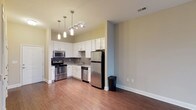 This single-floor space showcases a well-designed open-concept layout that connects the kitchen and living areas seamlessly. The kitchen is a standout feature, outfitted with stainless steel appliances including a refrigerator, stove, microwave, and dishwasher. White shaker-style cabinetry lines the walls, complemented by a tasteful tile backsplash that adds texture and visual interest. A deep sink sits beneath upper cabinets, maximizing functionality. Pendant lighting fixtures hang above, including a multi-bulb track light and individual drop pendants, providing both ambient and task lighting. The warm hardwood flooring extends throughout the open space, creating a cohesive and inviting atmosphere. Neutral-toned walls in soft gray and beige tones contribute to a clean, contemporary aesthetic. The adjacent living area benefits from the open sightlines, making the overall space feel airy and expansive. Elevated ceilings further enhance the sense of openness. The layout is practical and efficient, offering a modern urban living environment with quality finishes throughout.