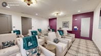 Located at 152 Market St Media in Pittsboro, NC, this single-floor living room is a thoughtfully designed communal screening and lounge space. The room is arranged in a theater-style layout, with rows of cream-upholstered reclining chairs dressed with teal throw blankets and decorative floral pillows, providing a cozy and inviting atmosphere for group gatherings. A plaid area rug anchors the seating arrangement and adds visual texture to the space.

The design is anchored by a bold plum-colored accent wall featuring elegant raised panel detailing, which frames a frosted-glass-panel door and serves as a dramatic focal point. Matching plum-painted cabinetry lines one wall, offering built-in storage and countertop surface space. A wall-mounted flat-screen television is positioned for optimal viewing from all seating areas, supported by in-wall speakers for enhanced audio quality.

Lighting is a blend of recessed ceiling lights and a stylish brass multi-globe flush-mount fixture that adds a warm, refined glow. A window along one wall allows natural light to filter into the space, brightening the neutral cream walls. Subtle gold accent tables are interspersed among the seating. The overall ambiance is polished yet comfortable, blending residential warmth with the functionality of a private screening room.