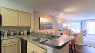 This single-floor residence showcases an open-concept design that seamlessly integrates the kitchen and living areas, creating a cohesive and functional living space. The kitchen is anchored by a peninsula-style counter finished in dark granite-style laminate, which extends outward to provide additional workspace and casual seating. A double stainless steel sink is built into the peninsula, adding convenience to the layout. Light-toned cabinetry with simple hardware lines the kitchen walls, offering ample storage. Track lighting mounted on the ceiling directs focused illumination across the kitchen area, enhancing both functionality and ambiance. The open sightline from the kitchen flows directly into the living area, where warm, neutral tones and soft furnishings create an inviting atmosphere. A large, colorful abstract artwork serves as a focal point on the shared wall between the two spaces, adding a vibrant touch. The living area is warmly lit by a floor lamp and benefits from natural light filtering through a window. Overall, the space presents a practical and comfortable layout with a casual, modern aesthetic suited for everyday living.
