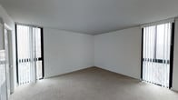 This single-floor space presents a clean, minimalist interior centered around an open room with a simple, functional layout. The walls are painted a crisp white throughout, creating a bright and airy atmosphere that is further enhanced by natural light streaming in through a large, floor-to-adjacent-ceiling window positioned along one wall. The window offers a view to the exterior, drawing daylight deep into the room and contributing to its open feel. Underfoot, neutral-toned wall-to-wall carpeting provides a soft, even surface across the space. The ceiling is smooth and flat, maintaining the room's uncluttered aesthetic. Electrical outlets are placed along the baseboards, offering practical utility points at convenient locations around the perimeter. The overall design is straightforward and versatile, with a neutral palette that serves as an adaptable backdrop suitable for a variety of uses. The absence of interior partitions in this area lends a sense of openness, while the combination of white walls, carpeted flooring, and generous natural light gives the space a clean, well-lit character.