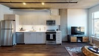 This contemporary urban loft showcases an industrial-modern aesthetic defined by exposed concrete ceilings with visible ductwork, rich dark hardwood flooring, and an open, airy layout that seamlessly connects the kitchen and living areas. The kitchen is well-appointed with sleek light-toned cabinetry, a white subway tile backsplash, stainless steel appliances including a gas range, built-in microwave, dishwasher, and full-size refrigerator, all set beneath the dramatic raw concrete ceiling. The living area benefits from large windows that frame expansive city and treetop views, allowing generous natural light to fill the space. A sliding barn-style door adds an industrial accent near the entryway. The bedroom offers a comfortable retreat with continued hardwood flooring, a raw concrete accent wall, and its own large window with picturesque views of the surrounding cityscape and tree canopy. A dedicated closet area provides practical storage. The bathroom features a clean, modern design with light wood cabinetry, a white countertop vanity, a backlit mirror, and a tub-shower combination. Throughout the unit, the interplay of raw concrete, warm wood tones, and bright white walls creates a refined yet industrial character that is both functional and visually distinctive.