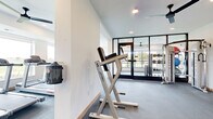 This contemporary fitness center is thoughtfully designed to support a full range of workout routines in a bright, clean environment. The space is organized into several distinct zones separated by sleek, floor-to-ceiling black-framed glass partitions, creating an open yet structured layout that maximizes functionality while maintaining visual connectivity throughout.

The cardio zone features elliptical machines and a stationary bike, positioned alongside expansive windows that offer pleasant outdoor views and fill the area with natural light. A dedicated strength training section includes Life Fitness equipment, a treadmill, weight bench, and a multi-tiered dumbbell rack stocked with a full range of free weights. An adjoining functional training area provides ample open floor space and houses a well-stocked accessory rack with stability balls, kettlebells, resistance bands, and yoga mats — ideal for stretching, core work, or group exercise.

Throughout the space, durable grey carpet flooring provides comfort and traction underfoot, while recessed ceiling lighting and multiple ceiling fans maintain a well-lit, comfortable atmosphere. Full-length mirrors in select zones enhance the sense of spaciousness and support proper form during exercise. Sanitizing stations are conveniently mounted on the wall. The overall design is clean, modern, and purposefully laid out to accommodate a variety of fitness needs.