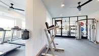 This contemporary fitness center is thoughtfully designed to support a full range of workout routines in a bright, clean environment. The space is organized into several distinct zones separated by sleek, floor-to-ceiling black-framed glass partitions, creating an open yet structured layout that maximizes functionality while maintaining visual connectivity throughout.

The cardio zone features elliptical machines and a stationary bike, positioned alongside expansive windows that offer pleasant outdoor views and fill the area with natural light. A dedicated strength training section includes Life Fitness equipment, a treadmill, weight bench, and a multi-tiered dumbbell rack stocked with a full range of free weights. An adjoining functional training area provides ample open floor space and houses a well-stocked accessory rack with stability balls, kettlebells, resistance bands, and yoga mats — ideal for stretching, core work, or group exercise.

Throughout the space, durable grey carpet flooring provides comfort and traction underfoot, while recessed ceiling lighting and multiple ceiling fans maintain a well-lit, comfortable atmosphere. Full-length mirrors in select zones enhance the sense of spaciousness and support proper form during exercise. Sanitizing stations are conveniently mounted on the wall. The overall design is clean, modern, and purposefully laid out to accommodate a variety of fitness needs.