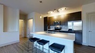 This single-floor residence showcases a thoughtfully designed open-concept layout that seamlessly connects the kitchen and living areas. The kitchen serves as the centerpiece of the space, anchored by a generous island topped with a crisp white quartz countertop that doubles as a breakfast bar, comfortably seating multiple guests on sleek upholstered stools. A deep double-basin sink with a modern arc faucet is integrated directly into the island for added convenience.

Dark espresso cabinetry lines the kitchen walls, providing rich contrast against the light surroundings, while a full suite of stainless steel appliances — including a French door refrigerator, electric range, and built-in microwave — delivers both function and a polished aesthetic. A tile backsplash adds texture behind the range, completing the kitchen's refined look.

Overhead, a combination of track lighting and elegant glass pendant lights illuminate the space with warm, layered light. Durable wood-look flooring flows throughout, grounding the open layout with a warm, cohesive feel. The living area transitions naturally from the kitchen, offering a versatile and welcoming environment. Clean wall lines, neutral tones, and contemporary hardware contribute to an overall ambiance that is modern, functional, and move-in ready.