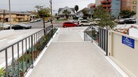 This single-floor building entrance presents a clean, modern design centered around accessibility and security. The approach to the entry is defined by a wide, gently sloped concrete ramp finished in a light aggregate surface, flanked on both sides by sleek metal railings. Flanking the ramp are neatly maintained landscaped planters featuring drought-tolerant succulents and ornamental shrubs, adding a touch of greenery to the otherwise minimalist exterior. The ramp leads to a secured entry vestibule, where a wall-mounted intercom and keypad system controls access to the building. Large floor-to-ceiling glass entry doors open into a bright lobby area, allowing natural light to filter through and creating a sense of openness at the threshold. Inside, light-toned floor tiles extend throughout the lobby, contributing to a clean and polished aesthetic. A built-in bank of individual mailboxes is conveniently positioned just inside the entrance, offering easy access for residents. Security cameras are noted at the entry points, reinforcing the building's commitment to safety. The surrounding streetscape, visible from the entry, includes residential homes and palm trees, suggesting a well-established urban neighborhood setting. Overall, the entrance conveys a contemporary, functional design with attention to accessibility and resident convenience.