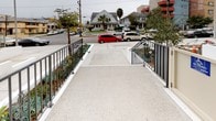 This single-floor building entrance presents a clean, modern design centered around accessibility and security. The approach to the entry is defined by a wide, gently sloped concrete ramp finished in a light aggregate surface, flanked on both sides by sleek metal railings. Flanking the ramp are neatly maintained landscaped planters featuring drought-tolerant succulents and ornamental shrubs, adding a touch of greenery to the otherwise minimalist exterior. The ramp leads to a secured entry vestibule, where a wall-mounted intercom and keypad system controls access to the building. Large floor-to-ceiling glass entry doors open into a bright lobby area, allowing natural light to filter through and creating a sense of openness at the threshold. Inside, light-toned floor tiles extend throughout the lobby, contributing to a clean and polished aesthetic. A built-in bank of individual mailboxes is conveniently positioned just inside the entrance, offering easy access for residents. Security cameras are noted at the entry points, reinforcing the building's commitment to safety. The surrounding streetscape, visible from the entry, includes residential homes and palm trees, suggesting a well-established urban neighborhood setting. Overall, the entrance conveys a contemporary, functional design with attention to accessibility and resident convenience.