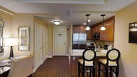 This single-floor space combines functionality and classic design in a welcoming layout. The kitchen serves as a central feature, equipped with rich medium-toned wood cabinetry, a stainless steel French door refrigerator, and a built-in microwave. Granite countertops extend into a raised breakfast bar, accommodating stylish black cane-back bar stools that add a refined touch. Elegant pendant lights hang above the bar, complementing the recessed and flush-mount ceiling fixtures that provide warm, consistent lighting throughout. A tile backsplash adds texture behind the cooking area, while a coffeemaker and small appliances suggest a fully functional setup. The flooring throughout is a rich dark hardwood, creating a cohesive and polished look. A wide hallway connects the kitchen area to additional rooms, lined with white-paneled doors and warm yellow-toned walls that lend the space a cozy, inviting ambiance. The entry door, featuring a keycard access panel, is visible at the end of the corridor, indicating a professionally managed property designed for comfortable, convenient living.