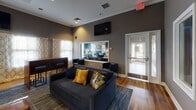 The Orchard Meadows community clubhouse and leasing office is a thoughtfully designed single-floor space that blends functionality with contemporary style. The interior is defined by a neutral palette of charcoal and warm taupe wall tones, accented by a bold teal feature wall and rich hardwood-style flooring that flows throughout the main areas, complemented by patterned area rugs that anchor each zone.

The central lounge area serves as the social hub of the space, furnished with plush dark gray sofas dressed in gold and navy accent pillows. A wall-mounted flat-screen television and a bar-height counter with leather stools provide additional gathering options. Four framed interior windows overlook the adjacent fitness center, offering a visual connection to the workout equipment within while maintaining a clean architectural detail.

A dedicated business center alcove, visible through a frosted-glass-paneled door, features computer workstations and professional seating. The leasing office, clearly branded with Orchard Meadows signage, provides a welcoming professional environment with workstations and natural light from exterior windows. Crown molding and recessed lighting add refined finishing touches throughout, while curated wall art contributes to the overall polished, community-oriented ambiance.